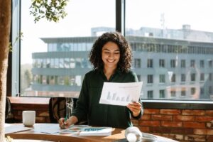 Compte Pro : cette banque pro vous offre 3 mois gratuits ! Smiling woman in a dark green shirt reviews charts at a sunlit wooden desk by a brick wall and large window.